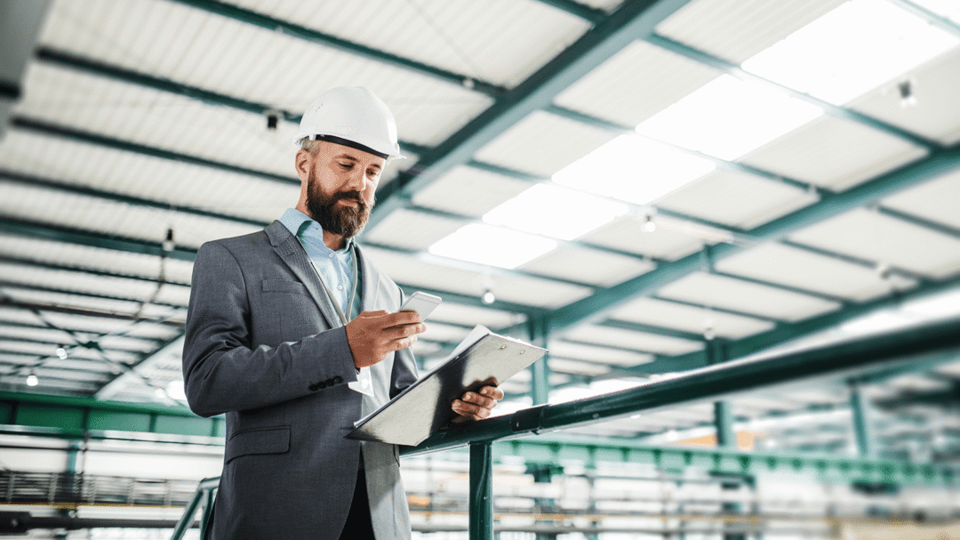 Engineer in a suit and hard hat reviewing plans on a clipboard inside a manufacturing facility